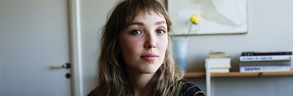 Ung kvinna som sitter i hemmiljö och tittar in i kameran.